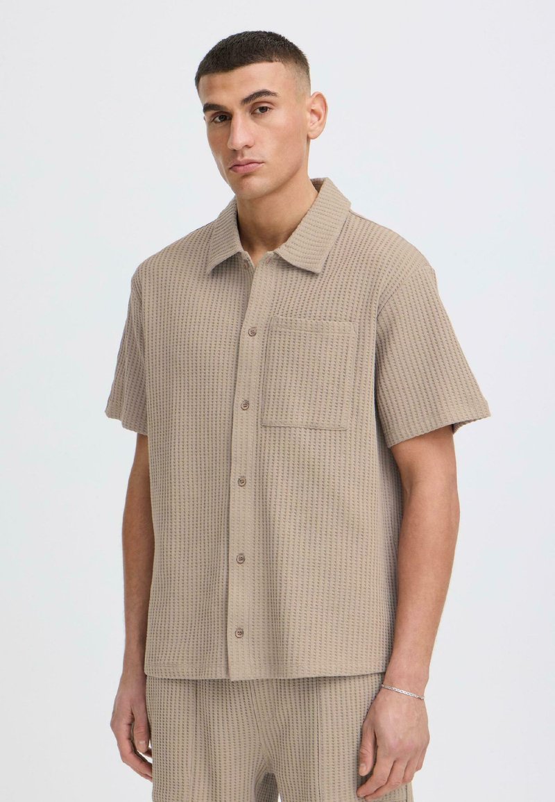 Homme portant une chemise beige à manches courtes texturée avec poche poitrine et un pantalon assorti, sur un fond uni.