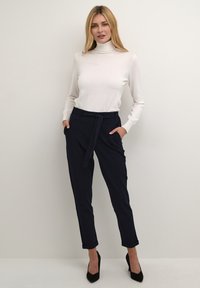 Pulover bej deschis cu guler înalt, asortat cu pantaloni bleumarin închiși, croiți, prevăzuți cu o curea după model, textură subtilă și manșete întoarse. Pantofi stiletto negri.