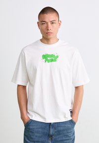 Mannlig modell med kort hår iført en hvit oversized T-skjorte med grønn "RIPNDIP PUMA" tekst og tegneserieikatter, sammen med blå jeans.