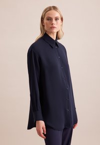 Camicia blu navy con bottoni in tessuto leggero, dotata di colletto, maniche lunghe e sottili dettagli con bottoni sui polsini e sul davanti.