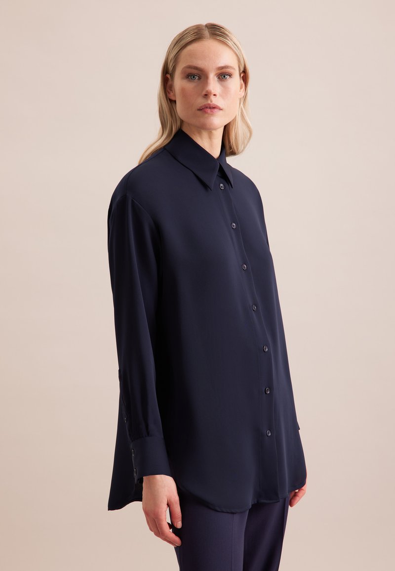 Camicia blu navy con bottoni in tessuto leggero, dotata di colletto, maniche lunghe e sottili dettagli con bottoni sui polsini e sul davanti.