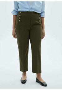Taille hoge olijfgroene broek met zes decoratieve zilveren knopen, gecombineerd met zwarte flats met perforaties, gedragen met een lichtblauw overhemd.