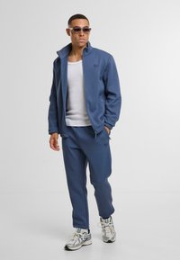 Zila sporta jaka un atbilstošas bikses no mīksta auduma, ar augstu apkakli un logotipa detalizāciju. Pabeigtas ar sudraba sporta apaviem.