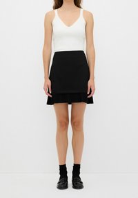 Černá plisovaná sukně s obtáhlým pasem, kombinovaná s bílým topem bez rukávů ve tvaru V. Oblečení má čistý, strukturovaný design. Černé ponožky a boty dotvářejí vzhled.