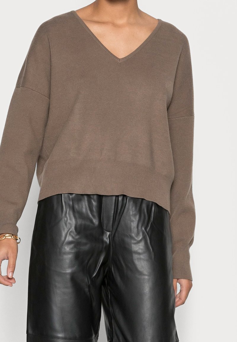 Personne portant un pull marron ample à col en V associé à un pantalon en cuir noir, debout devant un fond uni.