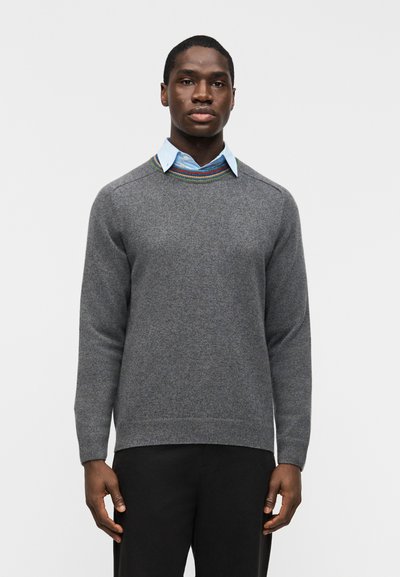 Homme portant un pull gris à col rond avec un col rayé multicolore par-dessus une chemise bleu clair boutonnée et un pantalon noir, debout.