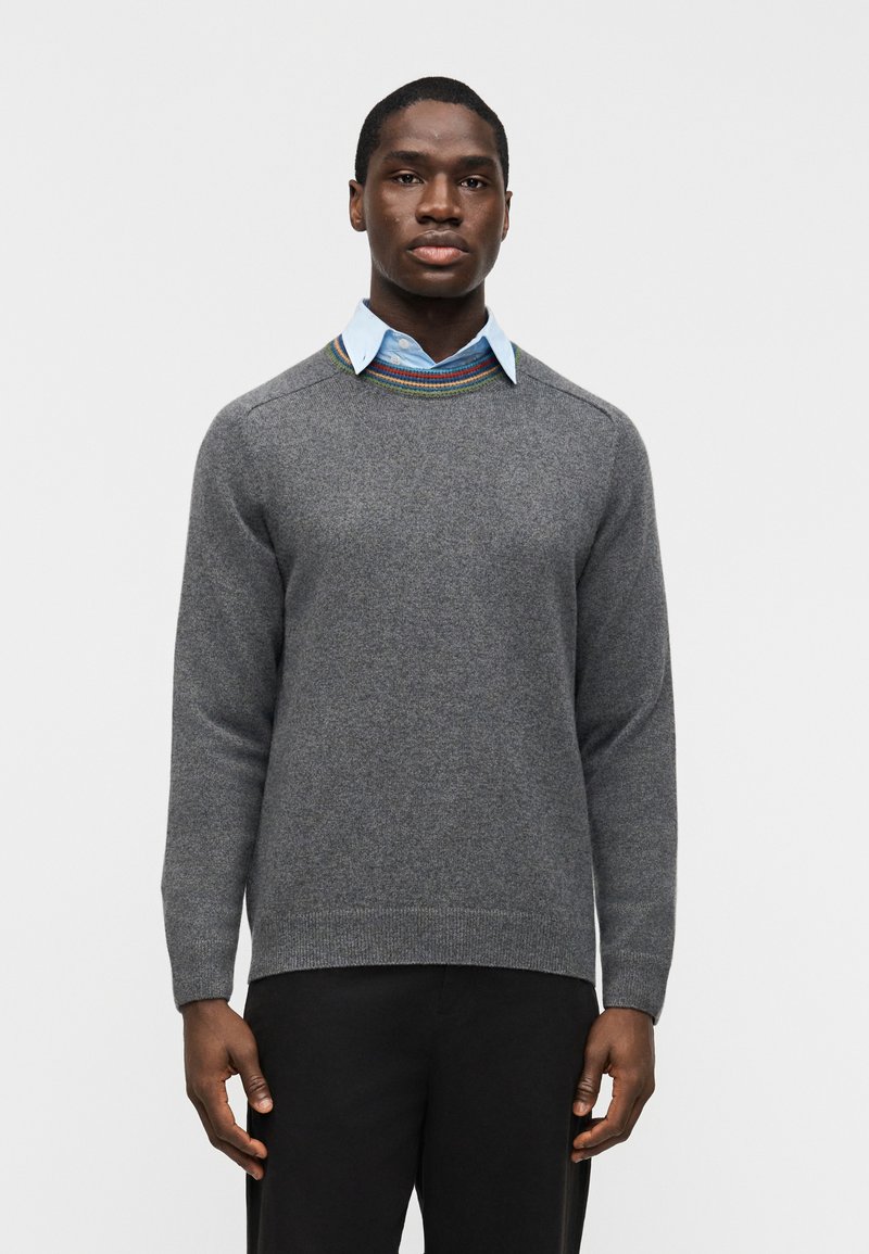 Homme portant un pull gris à col rond avec un col rayé multicolore par-dessus une chemise bleu clair boutonnée et un pantalon noir, debout.