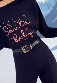 Fekete pulóver piros és zöld hímzett "Santa Baby" felirattal, flitterekkel és csillagokkal díszítve. Fekete krokodilmintás övvel párosítva.