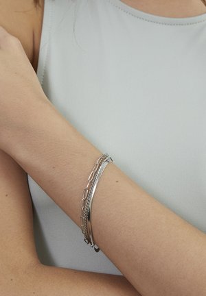 Drei silberne Armbänder in verschiedenen Designs: ein glattes Band, eine strukturierte Kette und ein Gliederarmband, getragen an einem gebräunten Handgelenk vor hellem Hintergrund.