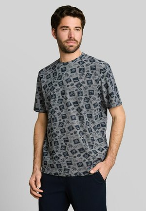 Man draagt een grijs t-shirt met zwarte tropische blad- en plantenpatronen, staat met één hand in zijn zak en kijkt iets naar opzij.