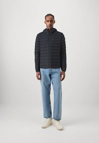 Veste noire matelassée avec fermeture éclair et capuche, associée à un jean droit bleu clair et des baskets blanches. Texture lisse avec un motif matelassé.