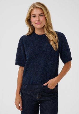 Femme blonde portant un pull court à manches courtes en maille duveteuse bleu foncé et un jean foncé, une main dans la poche, debout devant un fond clair uni.