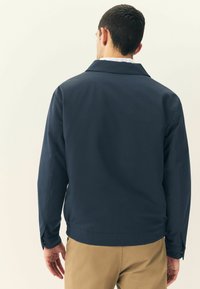 Giacca navy con una texture liscia, dotata di un colletto a pistagna e bottoni sui polsini. Indossata su pantaloni beige, vista da dietro.