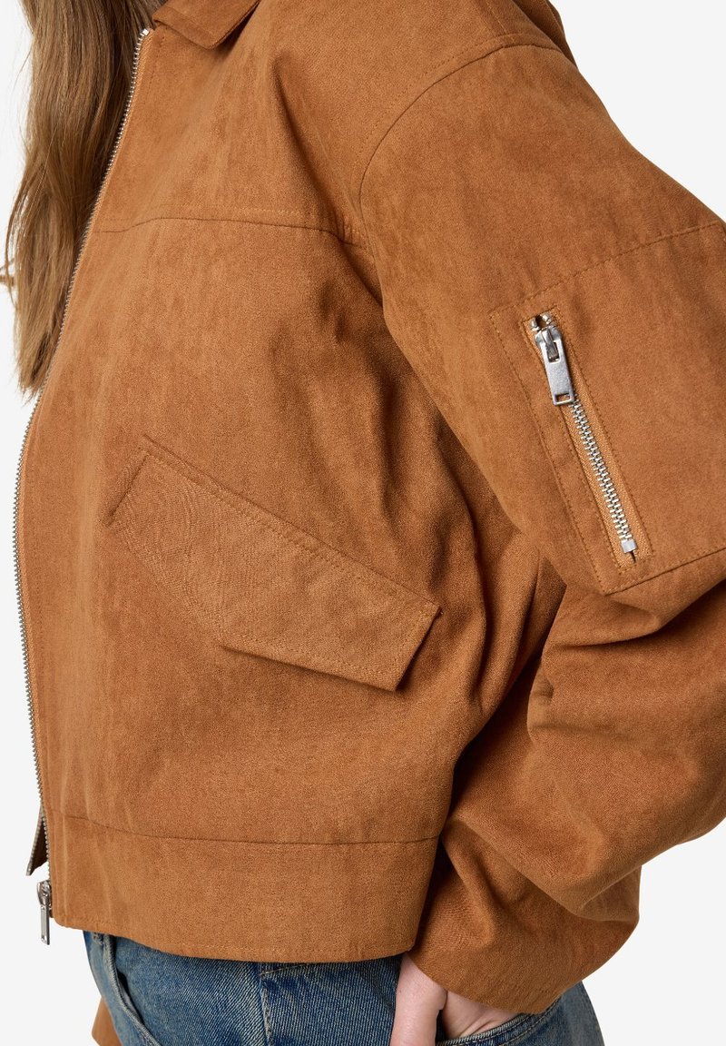 Veste cropped en daim marron avec une poche zippée sur la manche et une poche plate. Elle est dotée d'un col et d'une finition douce et texturée.