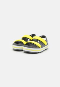 Mustat ja harmaat sandaalit, joissa on kaksi säädettävää keltaista hihnaa, uurrettu pinta ja valkoinen korostusraita pohjassa. Kevyt ja avara muotoilu.