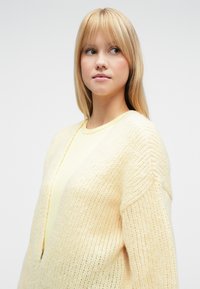 Šviesiai geltonas megztas kardiganas su riestu audiniu, atvira priekinė dalis, nuleistos pečių linijos ir ilgos rankovės, dėvimas ant geltonos palaidinės.