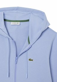 Sweatshirt zippé à capuche bleu clair avec des accents verts et un petit logo de crocodile brodé. Tissu doux avec une texture lisse. Coupe classique.