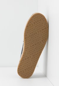 Semelle de chaussure avec motif de texte en relief répétitif "TOMMY HILFIGER" dans différentes orientations sur une surface en caoutchouc beige clair.