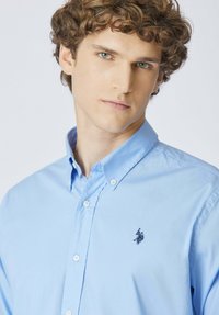 Giovane uomo con capelli castani ricci che indossa una camicia azzurra con bottoni e un logo ricamato scuro sul petto.