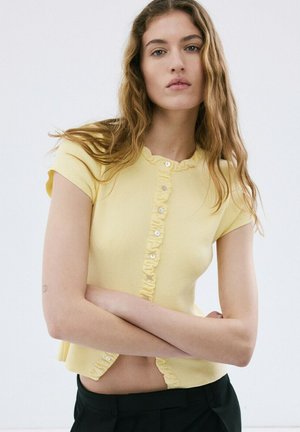 Joven con cabello largo y ondulado, vestida con una blusa amarilla de manga corta con botones y pantalones negros, con los brazos cruzados, sobre un fondo liso.