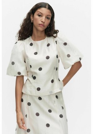 Vrouw die een witte outfit draagt met zwarte polkadots, opgeblazen mouwen en gouden oorringen tegen een effen witte achtergrond.