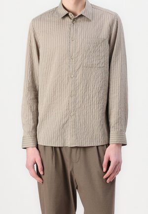 Mand iført beige skjorte med tynde lodrette striber og matchende lysebrune bukser mod en ensfarvet baggrund.