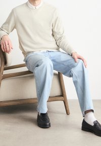 Béžová mikina s kulatým výstřihem, světle modré volné džíny, černé mokasíny, sedící na dřevěné židli. Jednoduchý design, bez vzorů nebo akcentů.