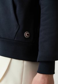 Mörkblå sweatshirt med ribbad mudd vid ärmslut och fåll, med en cirkulär logotypmärke i mörkrött på vänster sida, som visar ett stiliserat "C".