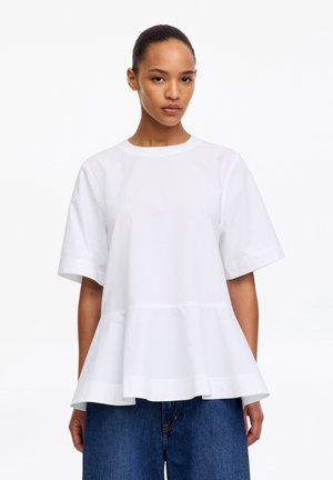 Femme portant un haut blanc ample à manches courtes avec un volant à la taille et un jean bleu, debout devant un fond blanc uni.