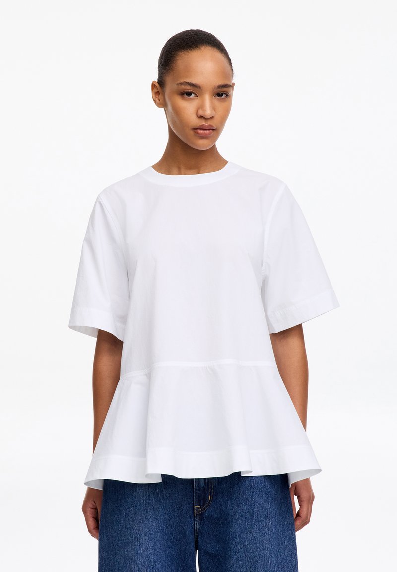 Femme portant un haut blanc ample à manches courtes avec un volant à la taille et un jean bleu, debout devant un fond blanc uni.