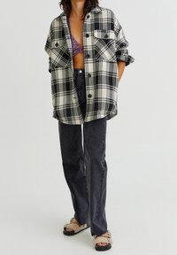 Zwart-witte geruite oversized hemdjacket met knopen, gedragen over een paarse top, gecombineerd met zwarte rechttoe-rechtaan jeans en beige sandalen.