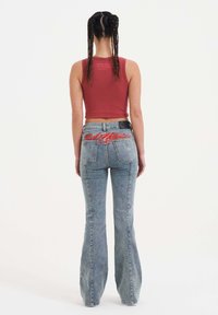 Top corto sin mangas rojo combinado con jeans acampanados de color azul claro. Los jeans presentan un logo bordado en rojo en la parte trasera y un ligero desvanecimiento.
