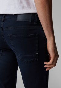 Man som bär mörkblå jeans med en bakficka och en läder BOSS-märkesetikett på midjebandet, stående mot en neutral bakgrund.