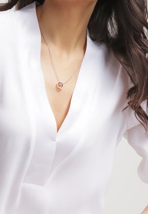 Vrouw die een witte blouse met V-hals draagt en een delicate roségouden halsketting met een cirkelvormige hanger die op haar borst rust.