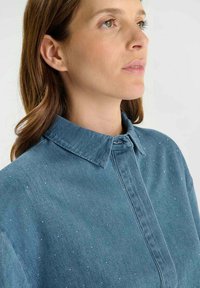 Denim overhemd in een blauwe tint, met een spread collar en subtiele kleine metalen studs door de stof. Knopening met knopen aan de nek.