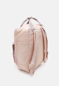 Plecak w kształcie sześcianu, wykonany z miękkiego różowego materiału, z regulowanymi paskami, uchwytami u góry oraz boczną kieszenią. Gładka tekstura z subtelnymi akcentami.