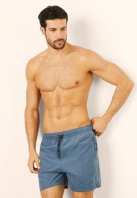 Modré plavky s elastickým pasem na šňůrku, vyrobené z lehkého materiálu. Mají volný střih a boční kapsy. Hladká textura.