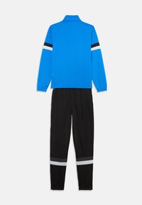 Plava majica s dugim rukavima s crnim i bijelim prugama na rukavima u kombinaciji s crnim hlačama koje imaju sive i bijele pruge iznad manžeta.