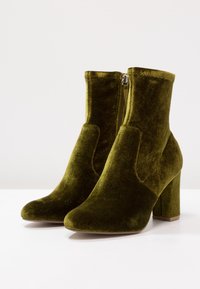 Une paire de bottes de cheville en velours vert olive avec des talons carrés et des zips sur les côtés, posées sur une surface blanche et un arrière-plan clair.