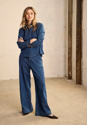 Žena stojaca so založenými rukami, oblečená v modrej denimovej košeli a širokých džínsach v priemyselnom štýle miestnosti.