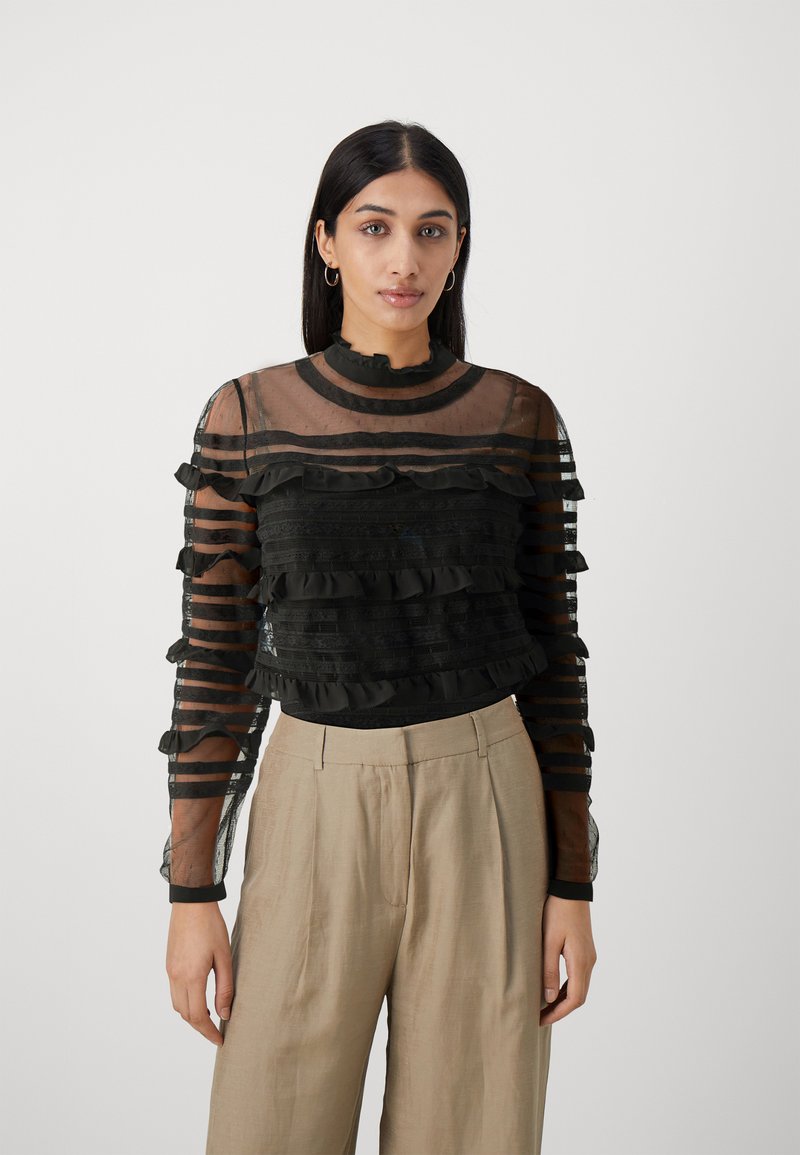 Top nero trasparente con dettagli a rouches orizzontali, colletto alto e maniche lunghe. Abbinato a pantaloni plissettati beige chiaro.
