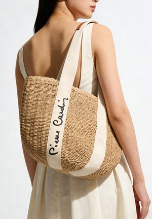 Femme portant une robe crème sans manches, portant un sac à bandoulière en paille tressée avec des sangles blanches et "Pierre Cardin" écrit sur une sangle.