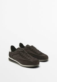 Sneaker in suede marrone con pannelli testurizzati, lacci piatti e un accentuazione bianca sul tallone. Sole in gomma con una lieve elevazione. Design minimalista.
