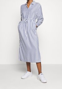 Gestreiftes Hemdkleid in Blau und Weiß, mit einem Bindegürtel, Knopfleiste, langen Ärmeln und seitlichen Taschen. Gestylt mit weißen Sneakers.