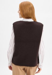 Frau mit langen roten Haaren, die eine dunkelbraune Fleeceweste über einem weißen Hemd mit roten Streifen und beigen Hosen trägt, von hinten gesehen.