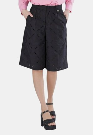 Donna con pantaloncini neri al ginocchio con ritagli a forma di diamante, abbinati a sandali platform neri e una maglia rosa a maniche lunghe.