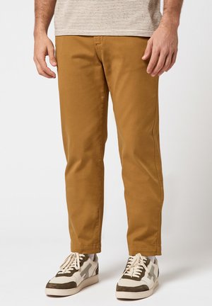 Homme portant un pantalon couleur moutarde, des baskets blanches avec des accents marron, et une chemise à manches courtes rayée, debout devant un fond uni.