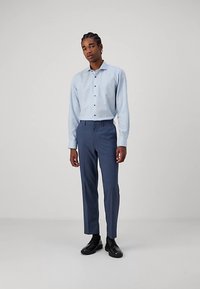 Chemise à motifs bleu clair avec boutons, pantalon ajusté bleu foncé et chaussures habillées noires, debout devant un fond uni.