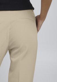 Beige, elegant kalhoty s hladkým povrchem a slim fit střihem. Mají zadní kapsu s čistým zakončením a decentním prošíváním.