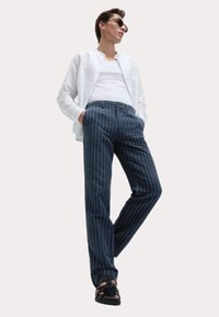 Pantalons marines à rayures fines avec une coupe ajustée, fabriqués en tissu léger. Associés à une chemise blanche et des sandales noires.